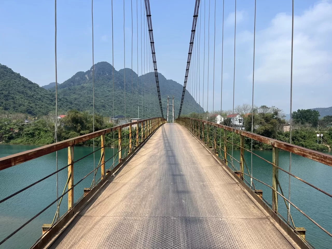 narrow road of a bridge running over a bright blue river with mountains peaking up in the background