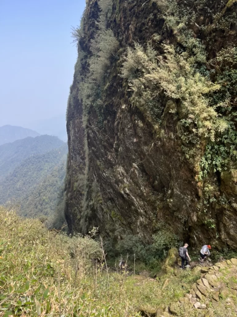 hikers climbing along a cliff face with an opening of a view to the surrounding landscape on its left