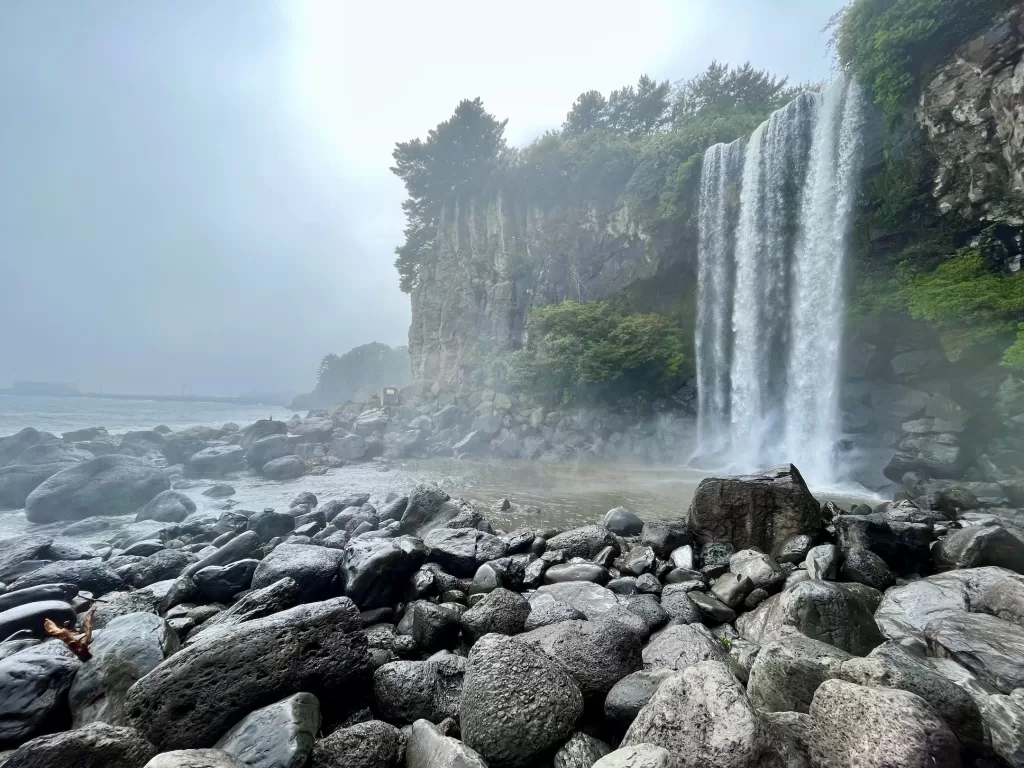 a collection of stones in front of a strong waterfall falling fast directly into the ocean with mist rising around the area