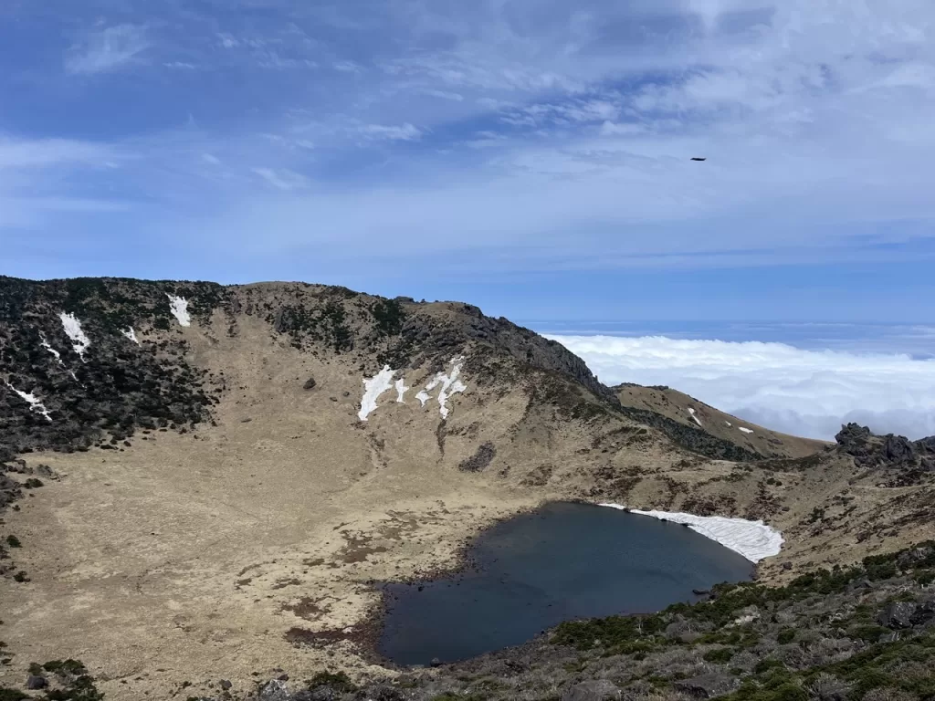 lake with snow around the edges at the top of a volcano with clouds visible underneath in the distance