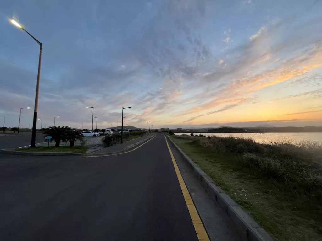 empty road with a beach on its right with pink clouds in the sky at sunset