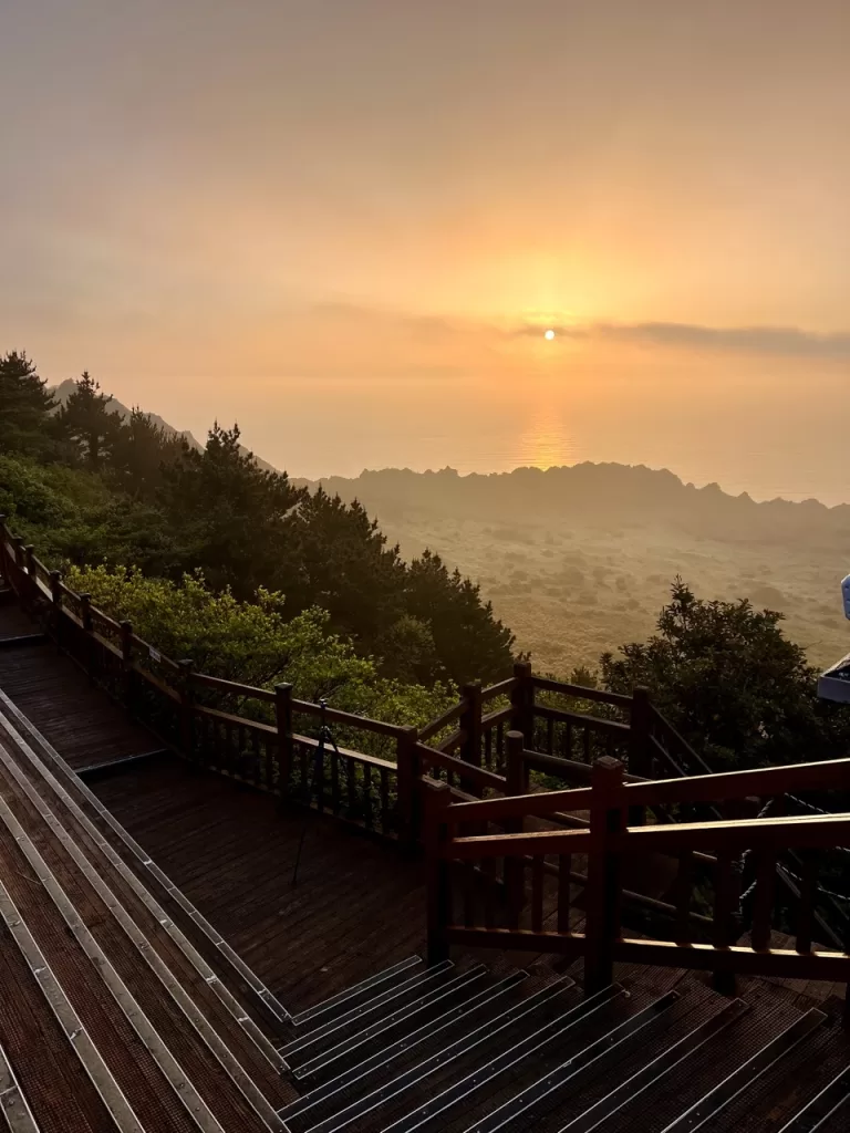 wooden deck overlooking a volcanic crater filled with lush trees with the sun rising over the sea in the background