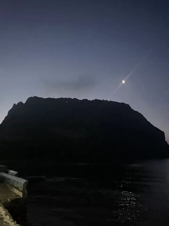 the silhouette of a small volcano in the dark skies of dawn with the moon on top