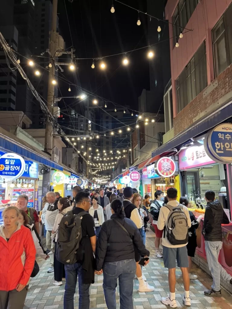 long narrow street food road at night with a lot of lights and filled with people