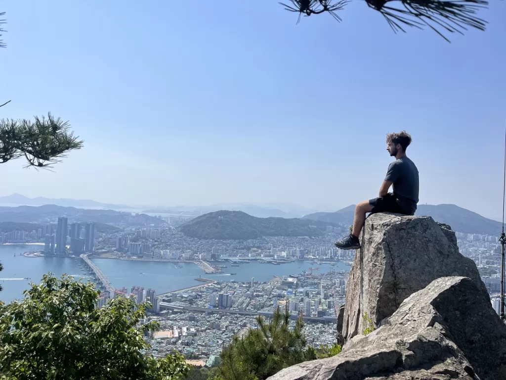 me sitting on a large rock overlooking a panoramic view of the sea and skyline of Busan
