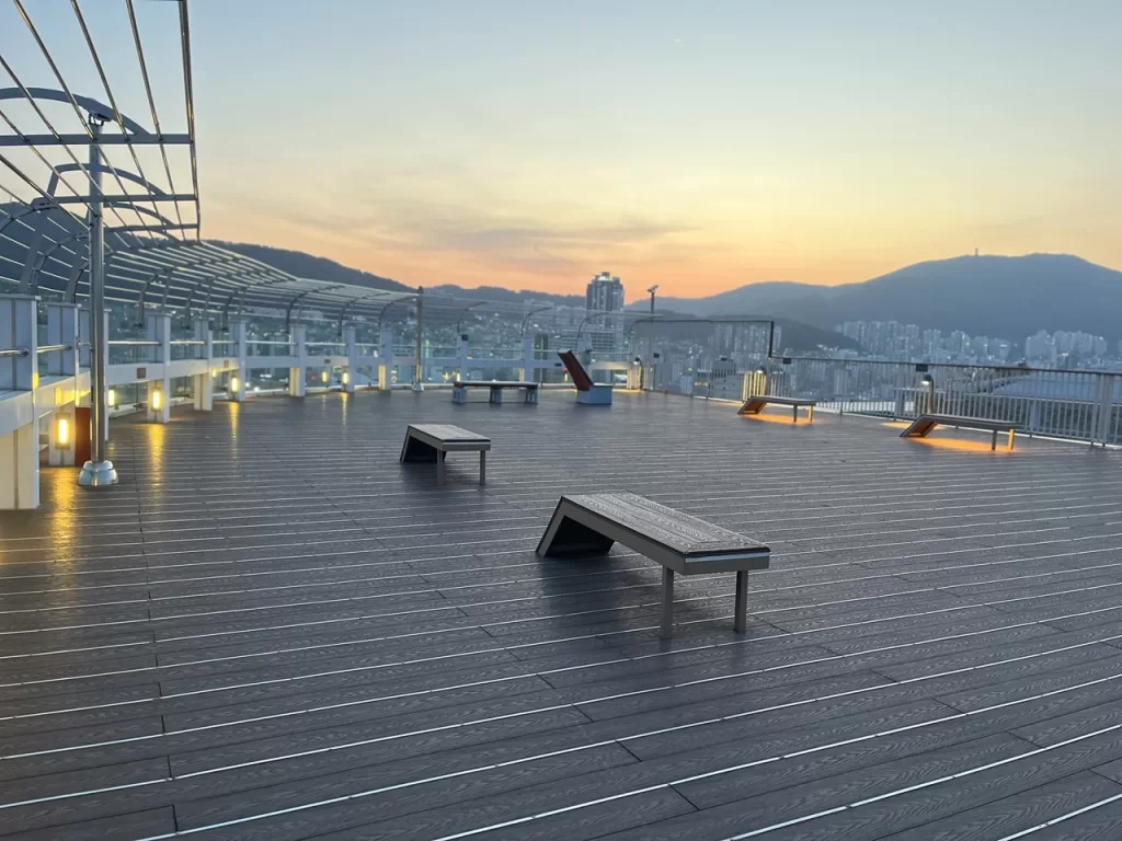 massive, empty rooftop with interspersed modern seats and glass barrier railing overlooking the sun setting over mountains over a city skyline