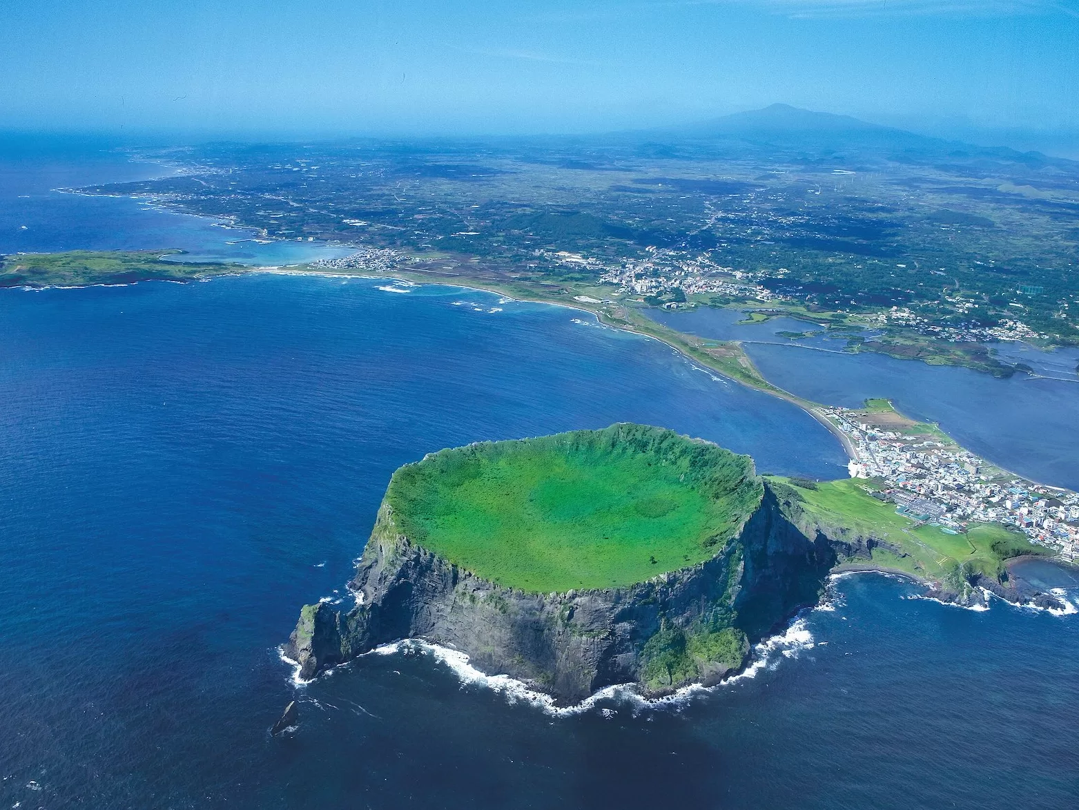 aerial view of a small volcanic island off a big island with a crater filled with bright green plants